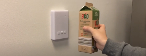 En hånd holder en mælkekarton op foran en hvid boks, der sidder på væggen.