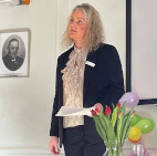Områdeleder Dorthe Nymann står i jakkesæt med et talepapir og en buket tulipaner i en vase foran sig.